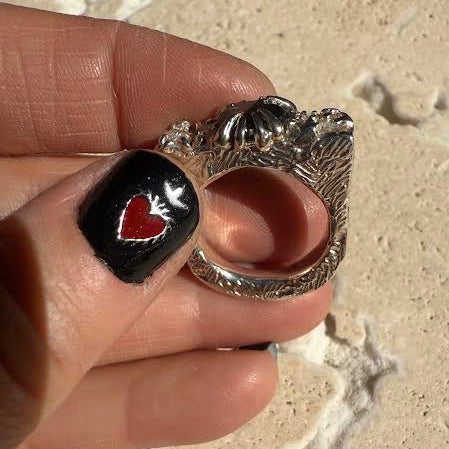 Silver ring with black stone and red heart design held by a hand on a textured surface.
