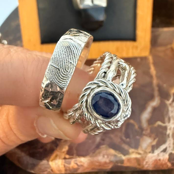 A hand holding two silver rings: one with a textured fingerprint pattern and the other featuring a central round dark blue gemstone surrounded by twisted silver details, with a jewelry display box blurred in the background.