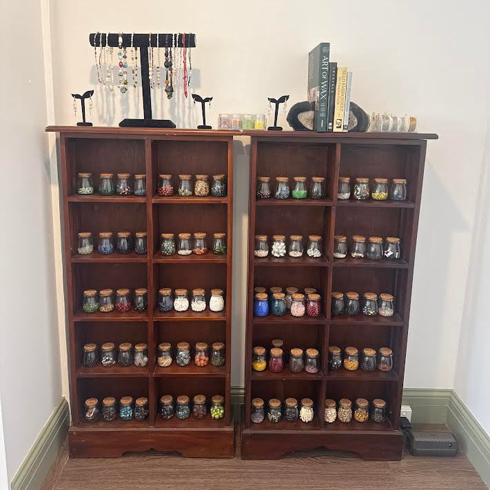 Two tall dark wood bookshelves filled with many small glass jars containing colorful items, set against a plain white wall on a wood floor. Jewelry stands and books sit on top of the shelves.
