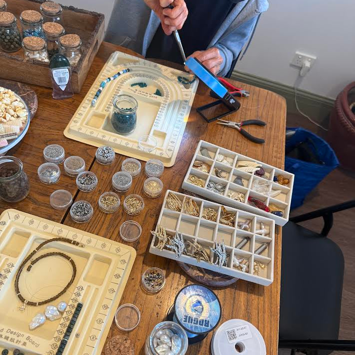 A person's hands and torso are visible as they work at a wooden table filled with jewelry-making supplies, including trays of beads, spools of thread, small jars, pliers, and a crafting mat.