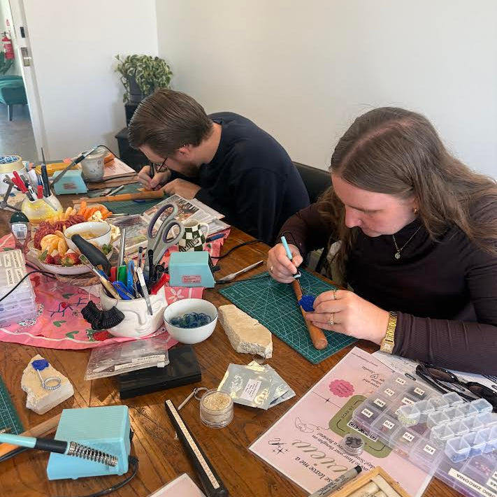 Two people seated at a wooden table in a home studio, working on jewelry-making projects. The table is cluttered with craft supplies including small bottles, brushes, tweezers, beads, and trays of small components.