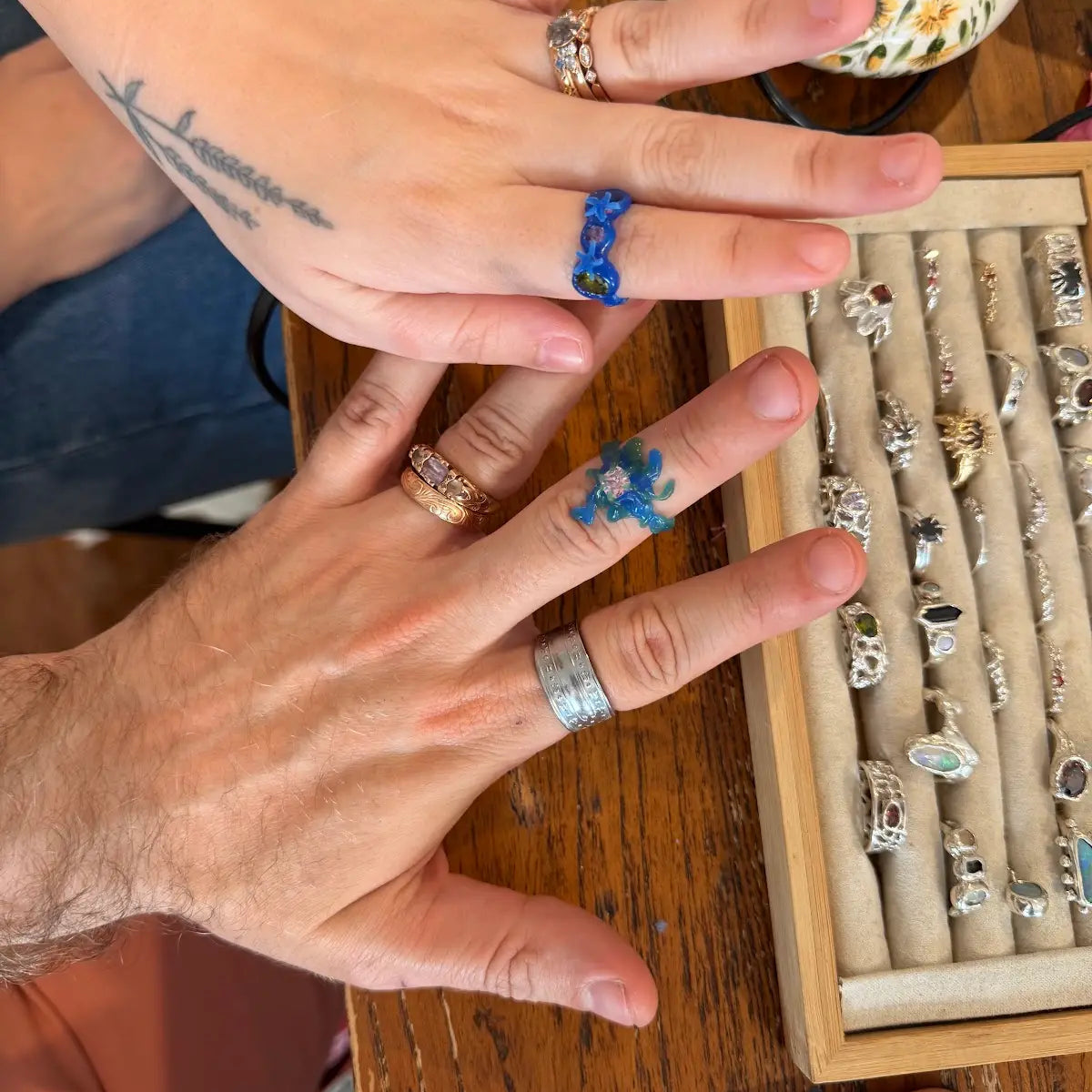 A close-up lifestyle shot of two hands displaying various handmade rings on a wooden table next to a ring display box. The hands feature temporary tattoos, and the background includes a crafting table and a smartphone.