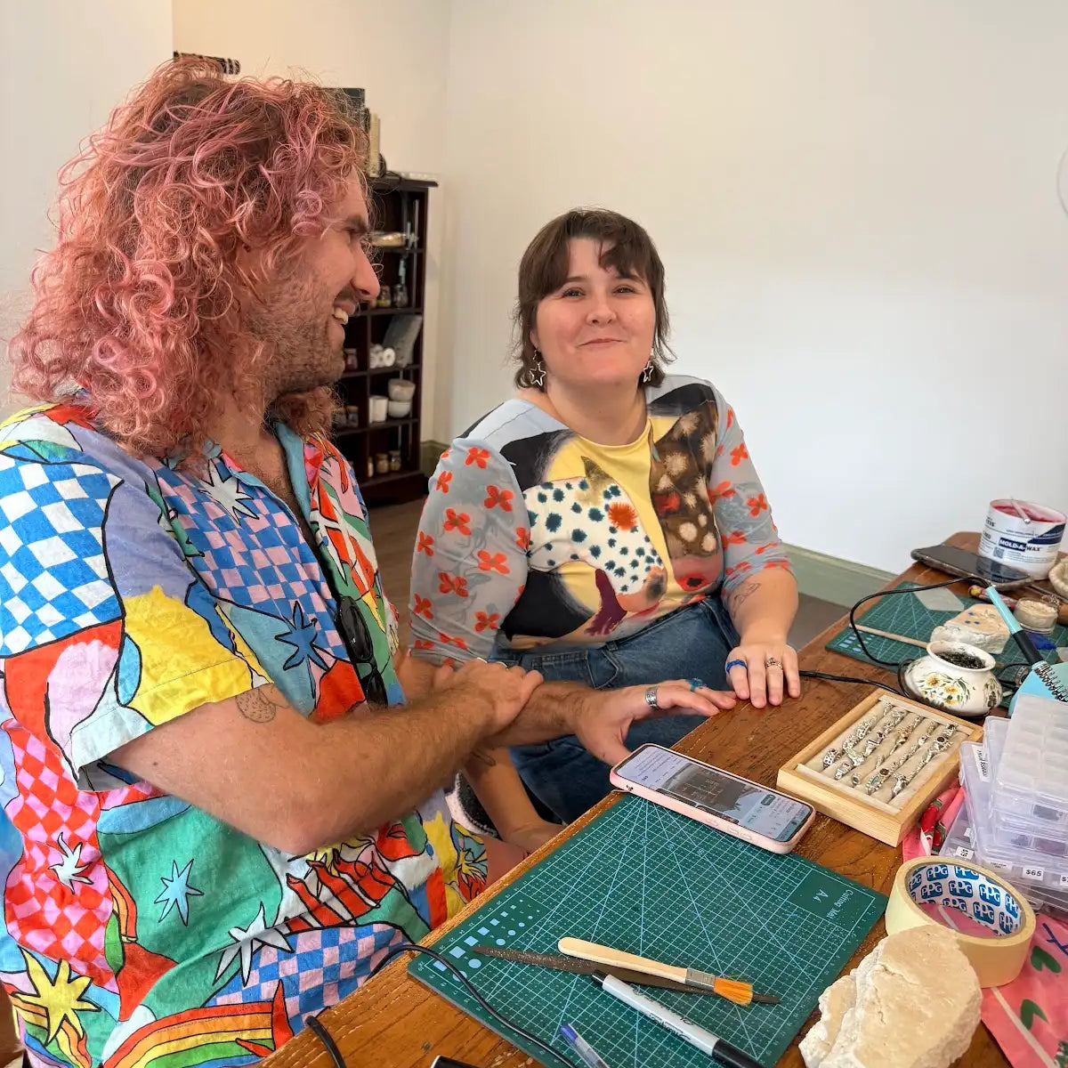Two people sitting at a wooden table in a craft studio, surrounded by cutting mats, stamps, and art supplies. The person on the left has curly pink hair and is wearing a colorful graphic t-shirt, while the person on the right is wearing a patterned cardigan over a yellow shirt.