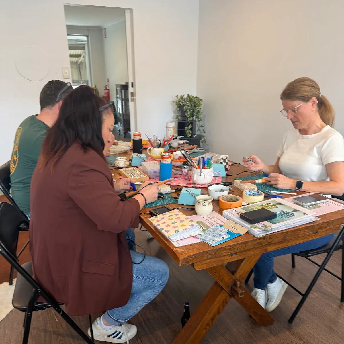 Three people seated around a wooden table in a home setting, engaged in a craft or painting activity with various art supplies, papers, and paintbrushes visible on the table.