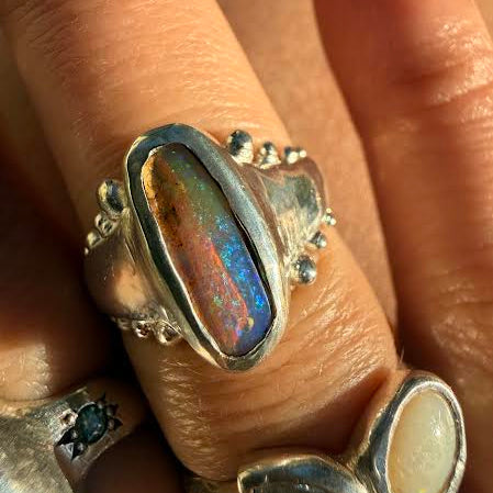 A close-up of a hand wearing a silver ring with a large iridescent opal cabochon, alongside other silver rings.