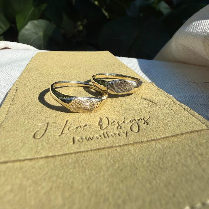 Two gold fingerprint ring bands resting on a tan suede jewellery card printed with 'J Line Designs Jewellery', set against a background of white fabric and dark green leaves.