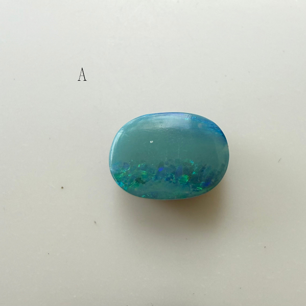 A polished opal cabochon doublet with a blue-green iridescent play-of-color, displayed on a plain off-white surface with a small letter 'A' printed nearby.