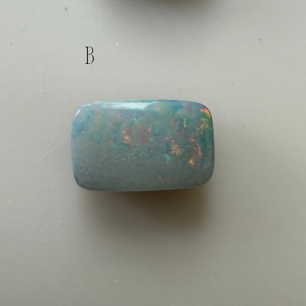 A polished rectangular cabochon OPAL with a light blue-green base and vivid iridescent flashes of orange, green, and yellow, shown on a plain light gray background with the letter 'B' printed above it.