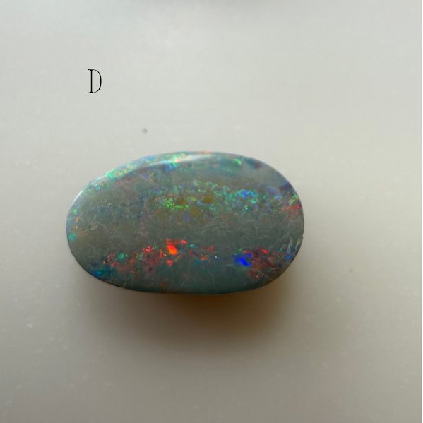 A polished oval-shaped opal stone with a gray-blue base and vibrant iridescent flashes of red, green, and blue, displayed on a light gray background with a small letter 'D' in the upper left.