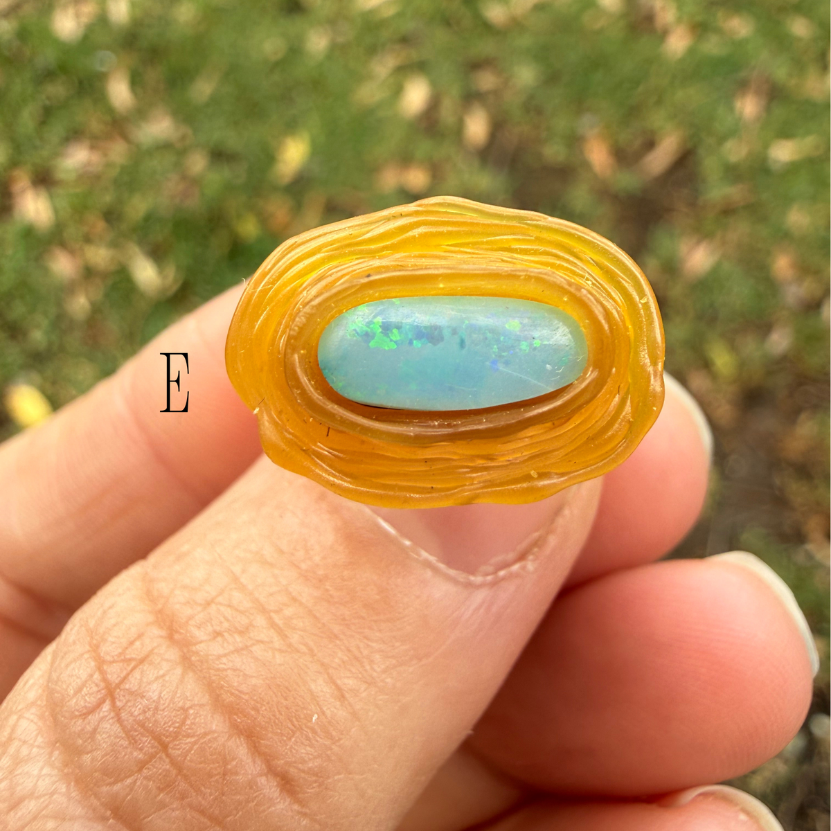 A hand holding a OPAL ring in wax form  and a light blue opal cabochon, set against a blurred green grass background.