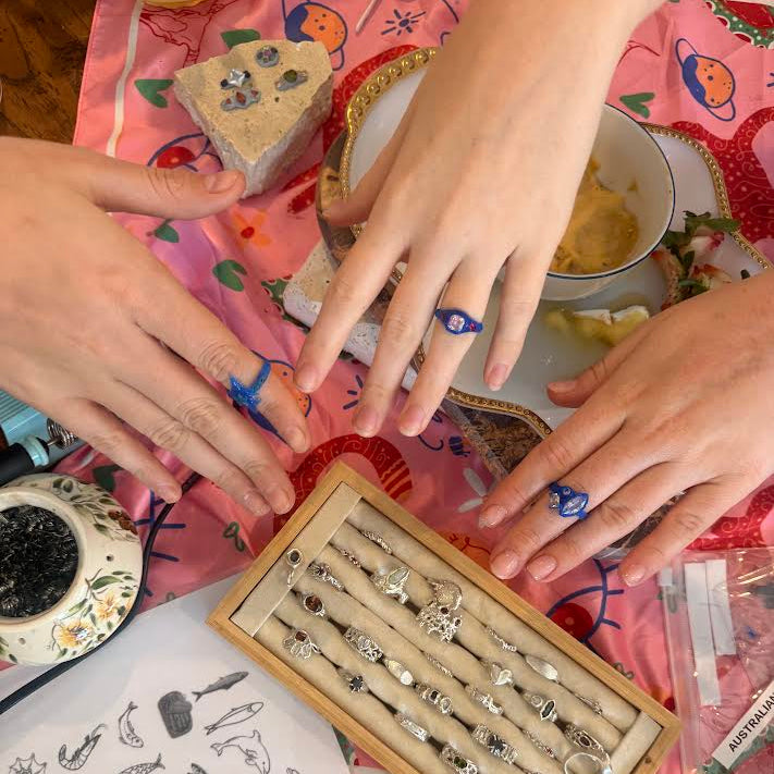 A top-down view of several hands wearing blue beaded rings, gathered around a wooden display box filled with silver rings on a pink patterned tablecloth.