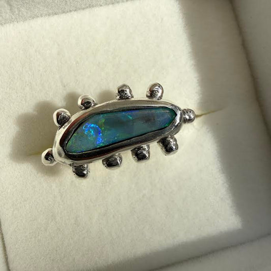 A sterling silver ring featuring a teardrop-shaped iridescent opal stone with blue, green, and orange flashes, displayed on a white velvet ring box.