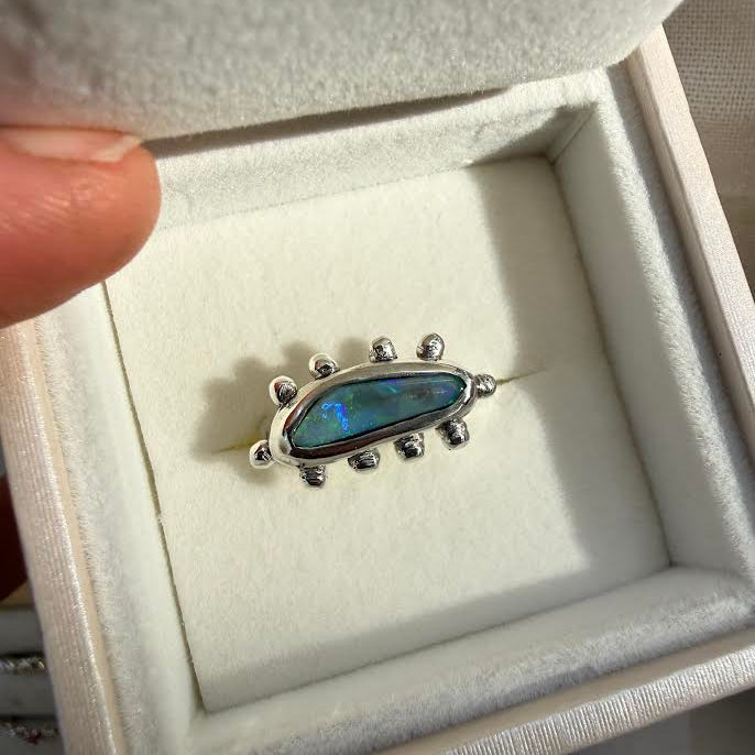 A silver ring with a central iridescent blue-green opal-like stone and small silver spikes, displayed inside an open cream-colored jewelry box. A finger is visible holding the box lid on the left.
