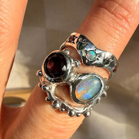 A close-up of a hand wearing a chunky, multi-stone silver ring. The ring features a large oval dark red gemstone, a smaller oval iridescent blue opal, and a textured silver band with small bead details and a small light blue stone.