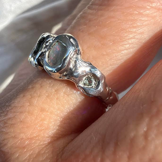 A close-up of a hand wearing a chunky, organic-shaped silver ring with a dark, polished central stone and a small clear accent stone on the side.