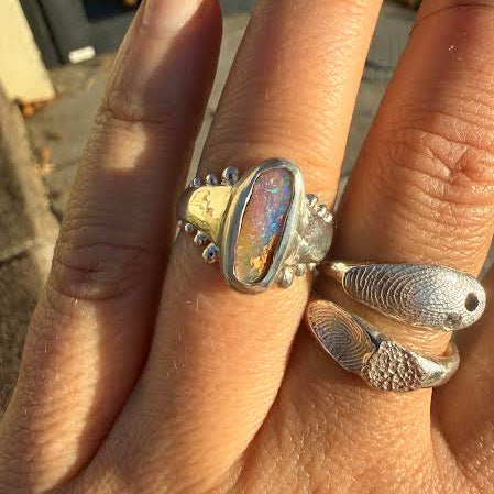 A close-up of a hand wearing two silver rings. The ring on the middle finger has a large oval iridescent stone, and the ring on the ring finger has two textured fish-shaped elements.