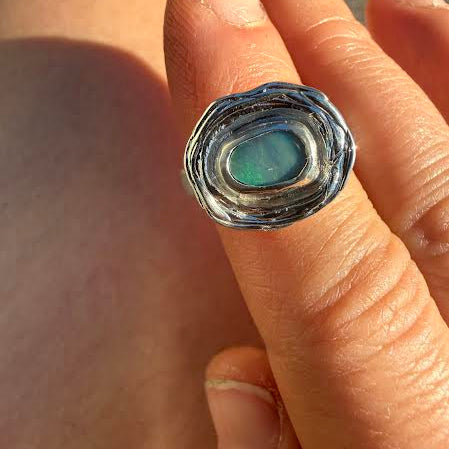 A close-up of a hand wearing a textured silver ring with a central iridescent green-blue stone.