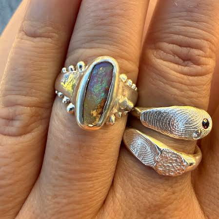 A close-up of a hand wearing a silver ring with a large iridescent oval opal-like stone and two textured fish-shaped rings stacked on the other fingers.