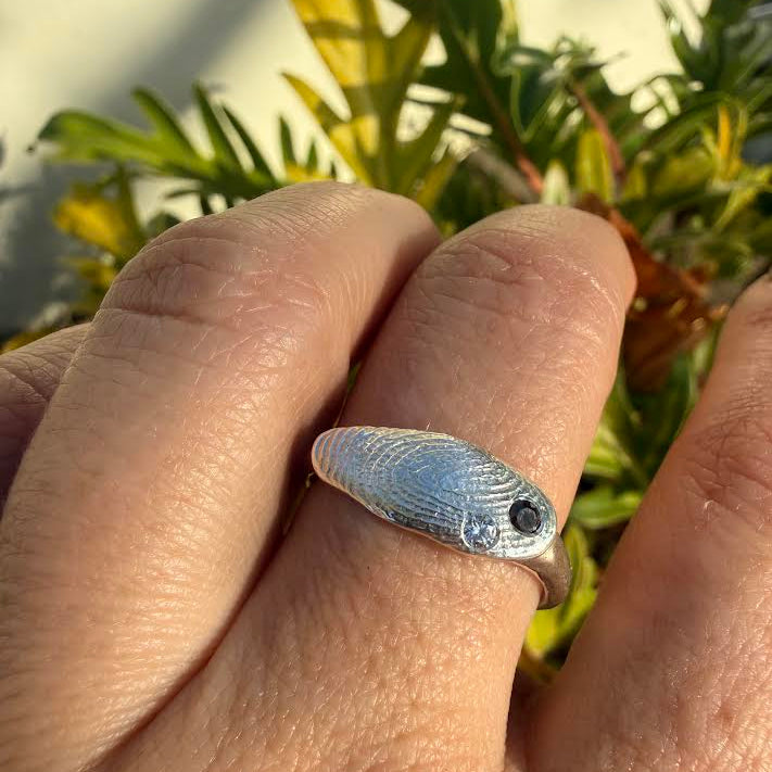 A silver ring shaped like a small fish with a textured scale pattern and a black eye, worn on a person's finger with green foliage in the background.