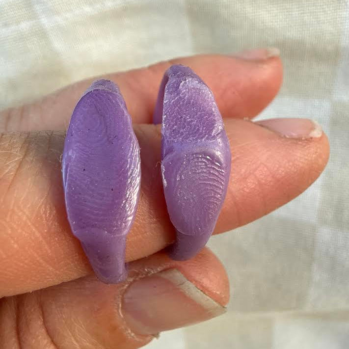 A close-up of a hand holding two small, purple, fingerprint-patterned items against a soft, light-colored fabric background.