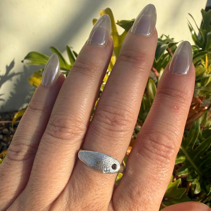 A silver ring shaped like a small fish with a textured surface and a tiny eye detail, worn on a hand with long, glossy nude manicured nails, set against a background of green foliage and a light wall.