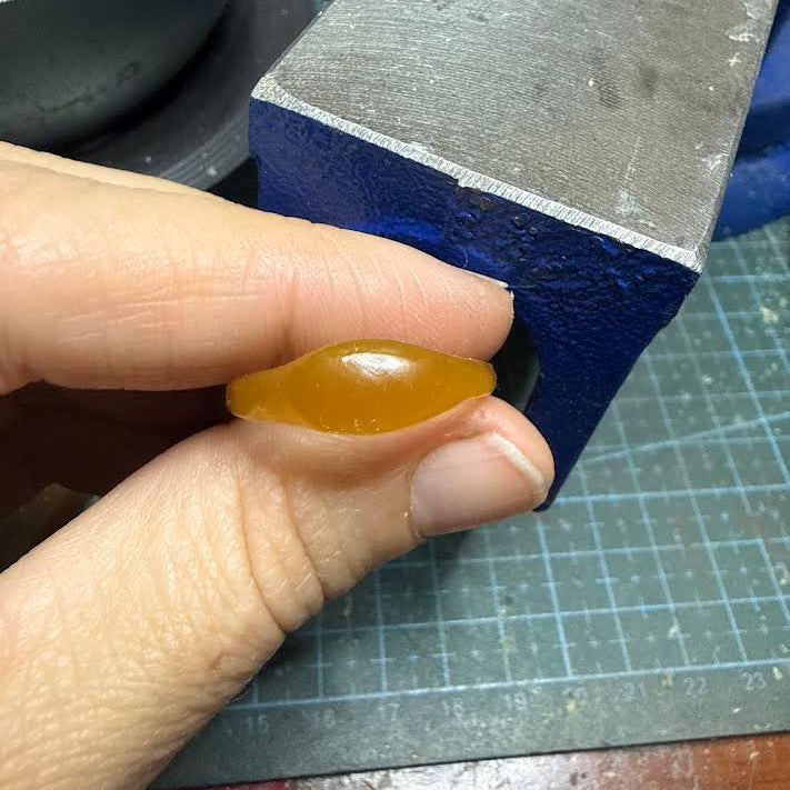 A hand holding a small, smooth, polished amber-orange stone or bead in front of a blue and gray metal bench block.