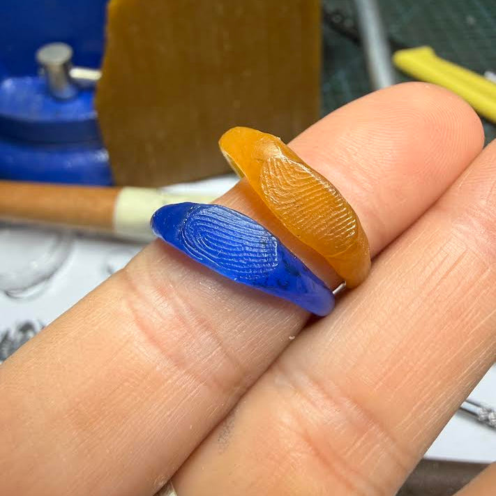 Two carved glass rings, one amber-orange and one translucent blue, stacked on a person's fingers in a workshop setting.