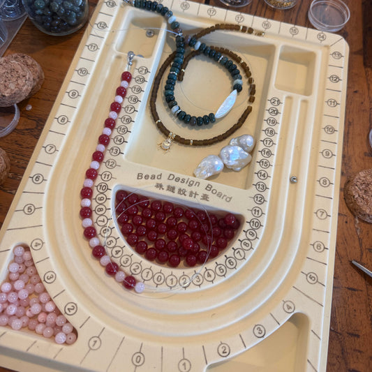 A beige bead design board laid on a wooden table, holding several beaded necklaces and strands of beads. Small clear jars containing loose beads are arranged around the board.